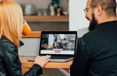 A couple sits at a desk, facing away from the camera, looking at an image of a home on the computer in front of them.