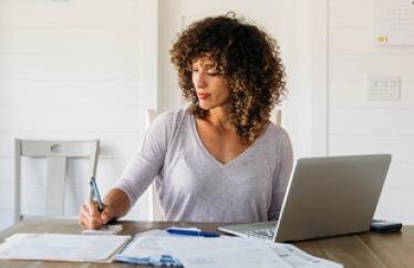 Woman sits at a desk in front of a computer; her right hand is writing on a piece of paper with a pen.