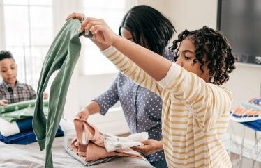 A young boy, middle-aged woman and young girl fold laundry on a bed. The young girl, closest to the camera, is holding green pants.