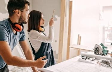 A man and a woman looking out a window with tools and paint around them