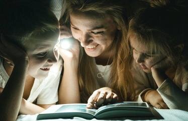 homeowner looking at books under the covers with their children while holding a flashlight